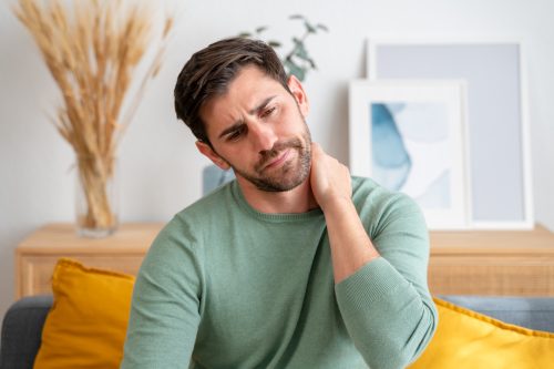 Young man feeling neck pain at home Young adult male grimaces in discomfort, rubbing his neck while sitting on a couch in a well-lit modern living room